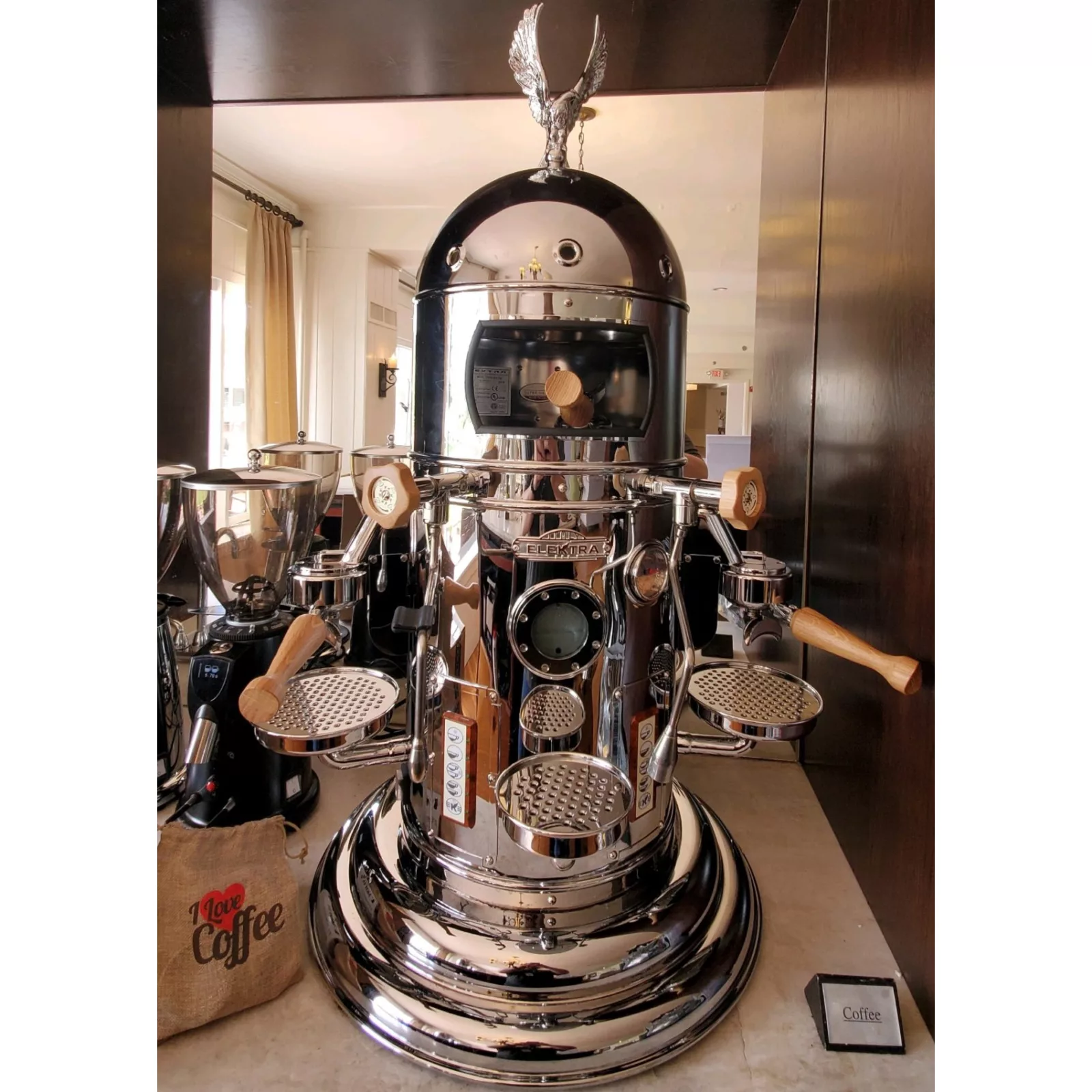 The Elektra Belle Epoque 2 Group Commercial Espresso Machine, featuring a dome top with an eagle ornament, wooden handles, and dual portafilters, sits among coffee accessories on a counter in a bright room. © 2025 1st-line.com