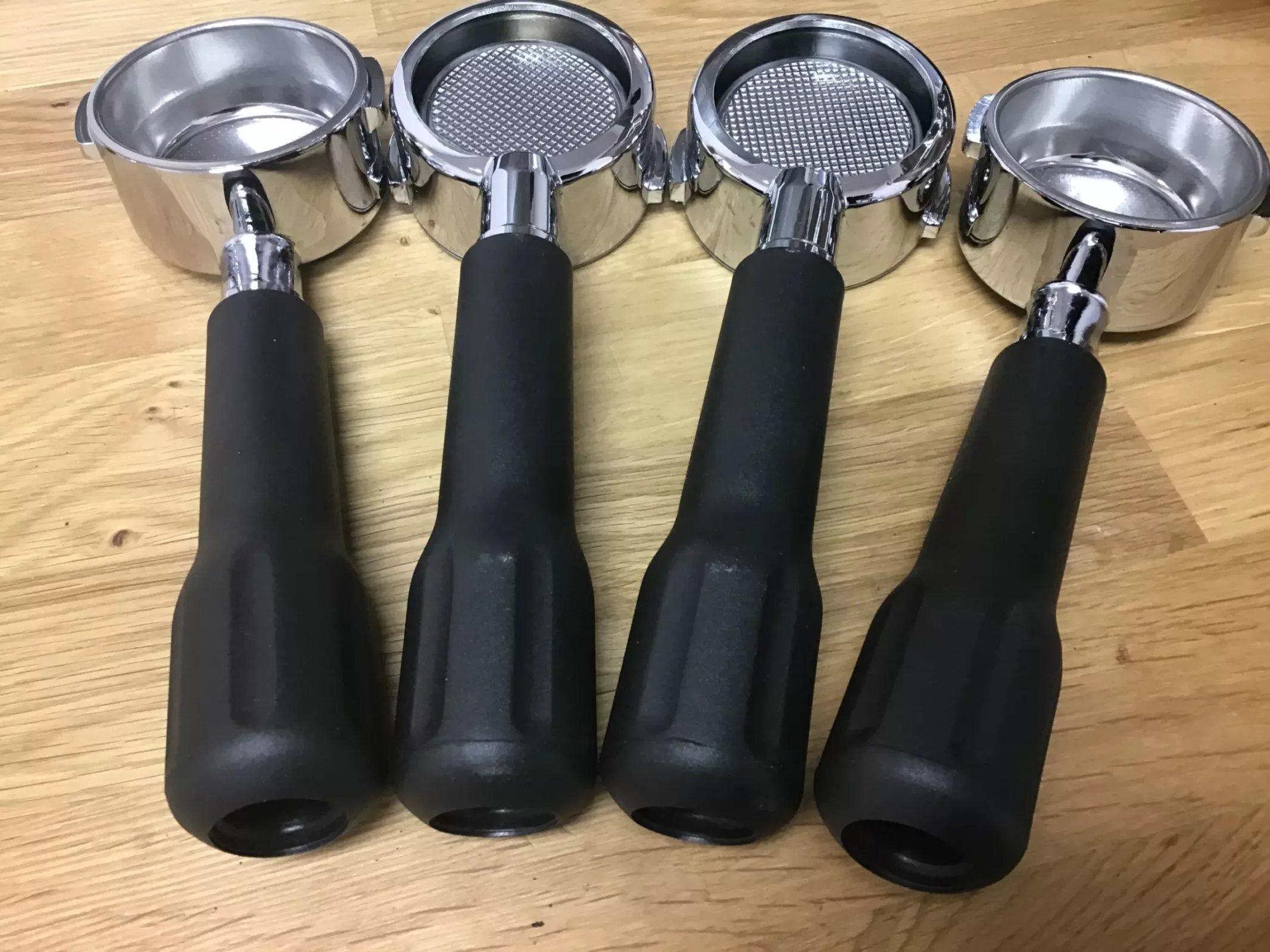 Four Lelit PLA570S 57mm bottomless portafilters with black handles are arranged side by side on a wooden surface, filter baskets facing up. © 2025 1st-line.com