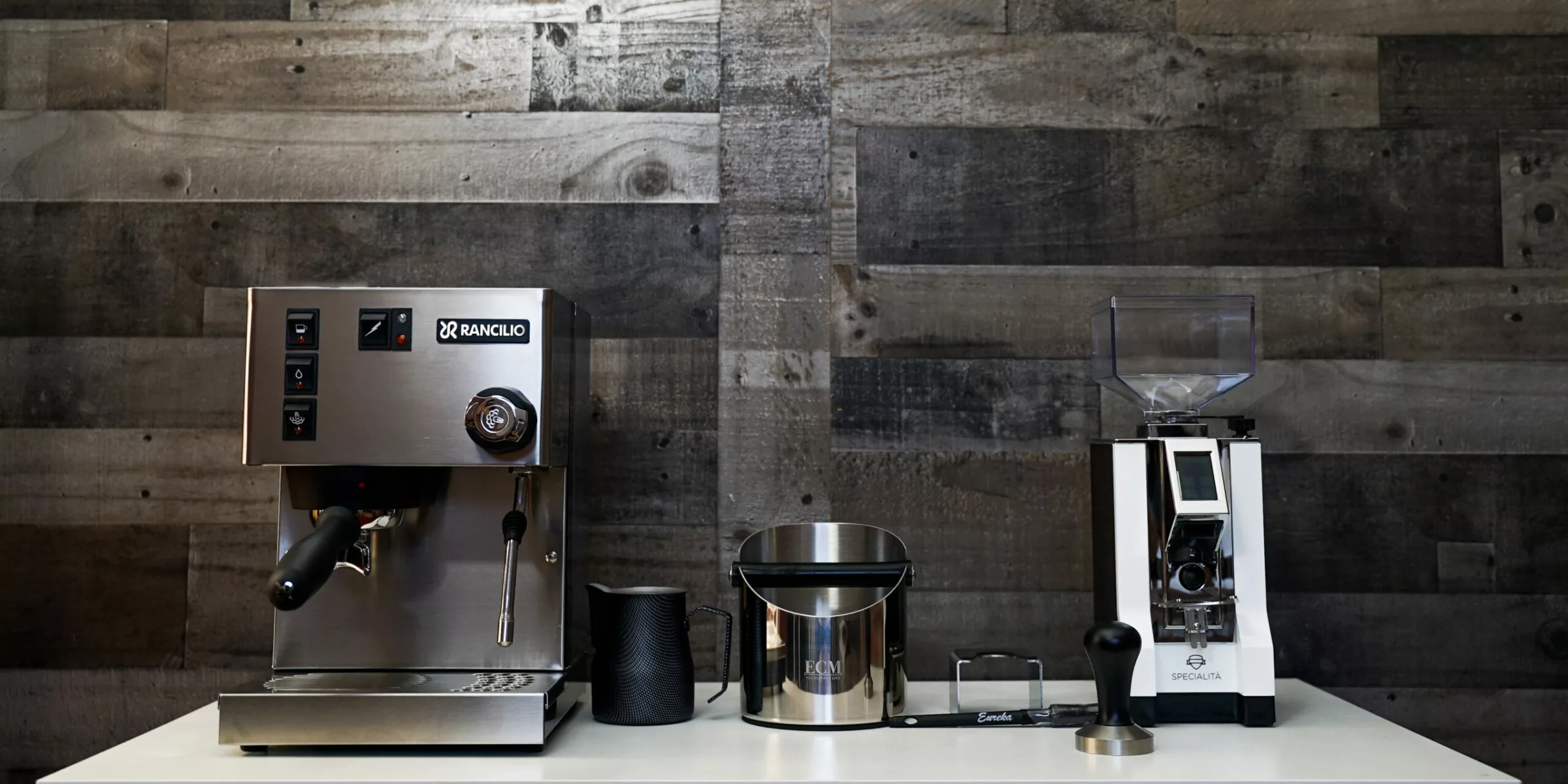 The Build My Rancilio Espresso Bar features a silver espresso machine and grinder on a white counter with coffee accessories like a knock box, tamper, and milk frothing pitcher, all neatly arranged against a rustic wooden wall. © 2025 1st-line.com
