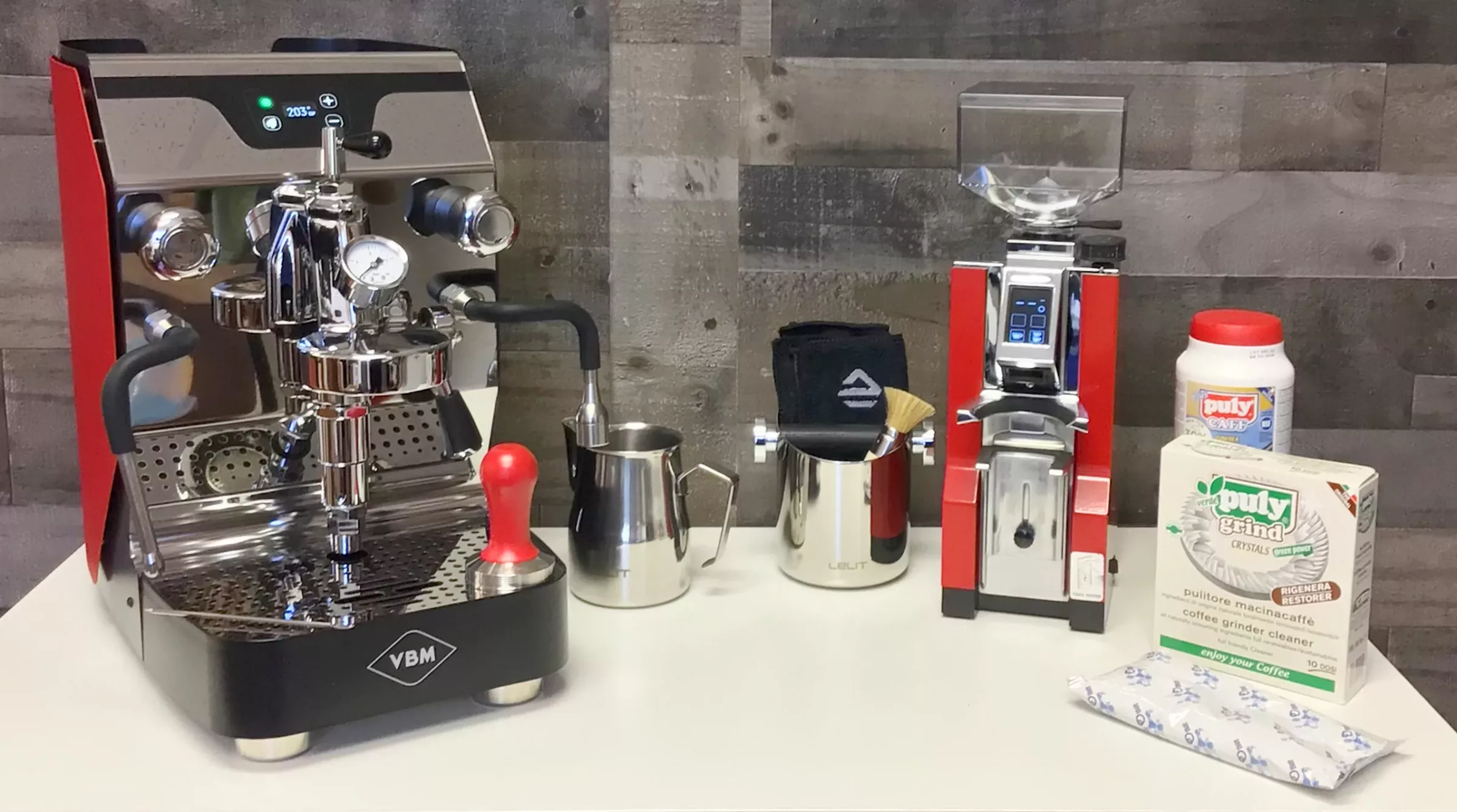 The Build My VBM Espresso Bar features an espresso machine, grinder, milk pitcher, and cleaning supplies on a white counter with wood panel wall and red accents—a stylish setup to inspire your own espresso bar at home. © 2025 1st-line.com
