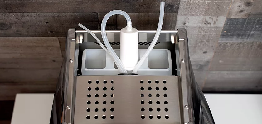 Top view of the DISCONTINUED La Nuova Era Cuadra HX Espresso Machine, showing its perforated metal panel, flexible tubes, and two white rectangular compartments on a white surface with a wood-paneled background. © 2025 1st-line.com