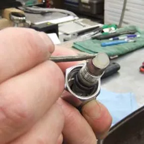 A close-up of a hand holding a spark plug while cleaning its electrode with a small tool in a workshop, similar to the careful precision needed in steam valve gasket repair. © 2025 1st-line.com
