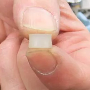 A close-up of a person holding a small translucent cylindrical object—ideal as a VBM Valve Gasket—between their thumb and index finger. © 2025 1st-line.com