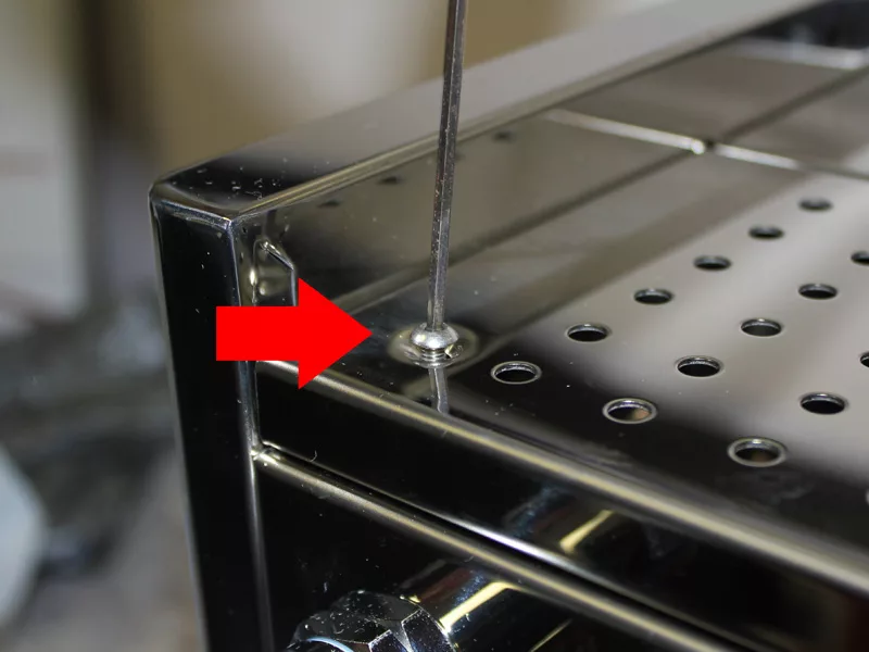 A close-up of a shiny metal appliance shows a red arrow pointing to a screw being tightened or loosened with a tool, an essential step in Water Tank Replacement. The surface features holes and a reflective finish. © 2025 1st-line.com