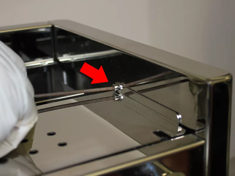 A close-up of a shiny metal appliance with a red arrow pointing to a small screw or connector on the water tank manifold, indicating a specific part for assembly or adjustment. A gloved hand is partially visible on the left side. © 2025 1st-line.com