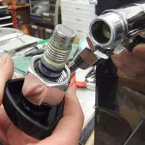 A close-up of hands assembling or repairing a metal plumbing fitting, possibly a faucet, with tools and various items visible in the background, highlighting a VBM repair or replacement of a hot water valve gasket. © 2025 1st-line.com