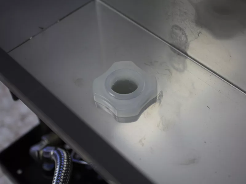 A close-up view of a white plastic hexagonal nut or cap inside a metal enclosure, part of a water tank manifold, with reflective surfaces and cables visible in the bottom left corner. © 2025 1st-line.com