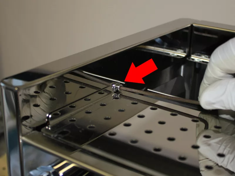 A gloved hand adjusts a shiny metal tray with holes, which is part of the water tank manifold, while a red arrow points to a small latch or tab on the tray’s surface. © 2025 1st-line.com