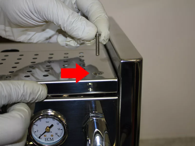 A gloved hand holds a screw above a metal espresso machine, prepping for a water tank replacement. Another hand points to the screw hole on top, highlighted by a large red arrow. A pressure gauge is visible on the front panel. © 2025 1st-line.com