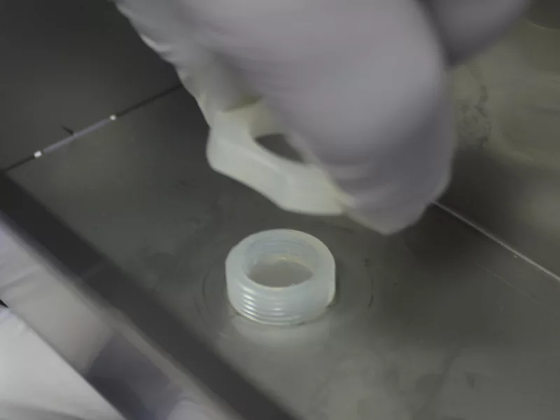 A gloved hand holds a white plastic ring above a matching threaded plastic fitting on a metallic surface, possibly preparing for water tank repair or replacing water tank components by securing the ring onto the fitting. © 2025 1st-line.com