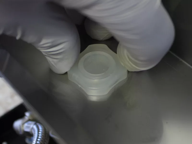 A gloved hand places or adjusts a translucent plastic cap inside a metallic container, suggesting a laboratory setting—possibly during a water tank replacement or while working with a water tank manifold. © 2025 1st-line.com