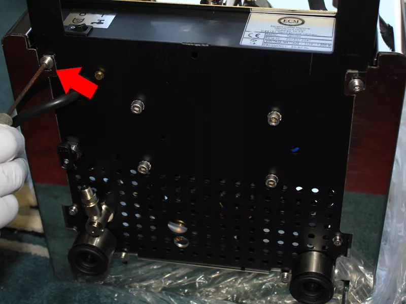 A gloved hand points with a screwdriver at a screw on the back panel of a black machine, indicated by a large red arrow—part of the process for replacing water tank components during water tank repair. © 2025 1st-line.com