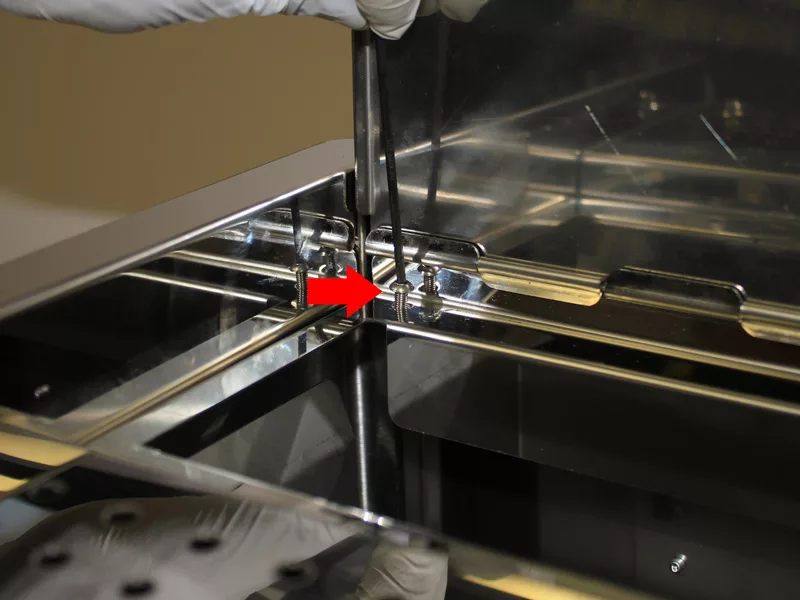A gloved hand uses tweezers to adjust a small metal component inside a reflective water tank manifold enclosure, possibly during a water tank repair. A red arrow indicates the precise area being worked on. © 2025 1st-line.com