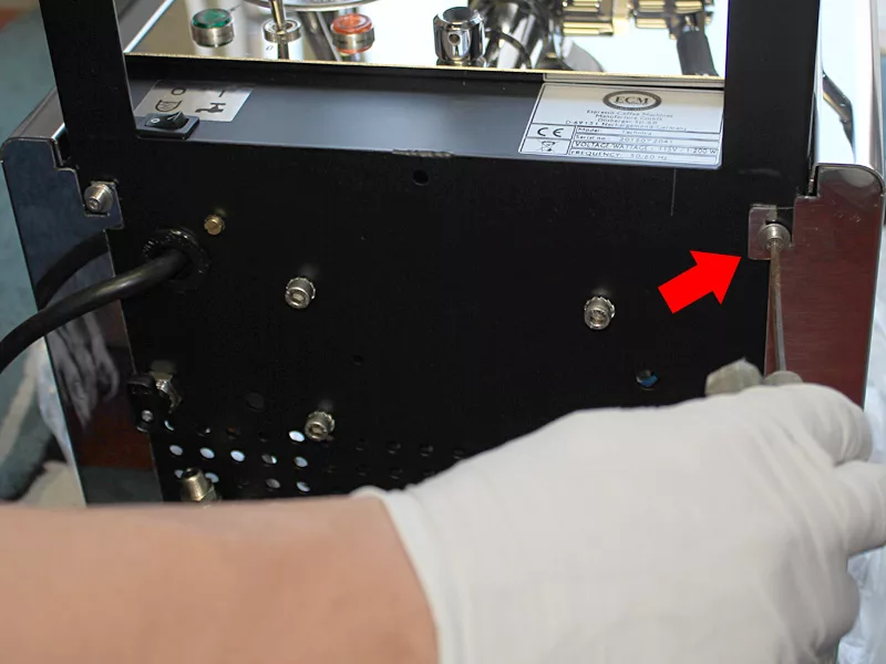 A person wearing a white glove uses a tool to unscrew a bolt on the side of a black electronic device, possibly as part of Water Tank Repair, with a red arrow pointing to the bolt. © 2025 1st-line.com