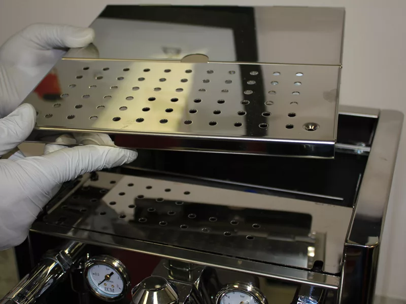 A person wearing white gloves lifts a perforated metal tray from the top of a stainless steel espresso machine, preparing for a water tank repair. Two pressure gauges are visible on the front of the machine. © 2025 1st-line.com