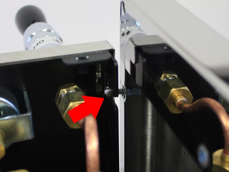 Close-up of a metal device in a water tank manifold, with a red arrow pointing to a small black screw between two brass fittings and copper pipes. These components are mounted on a black and silver frame. © 2025 1st-line.com