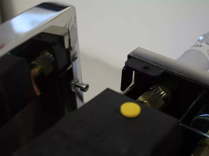 Close-up of a metal screw partially inserted into a hole between two mechanical components, likely part of a Water Tank Manifold, with one chrome finish and the other black plastic casing featuring a yellow button. © 2025 1st-line.com