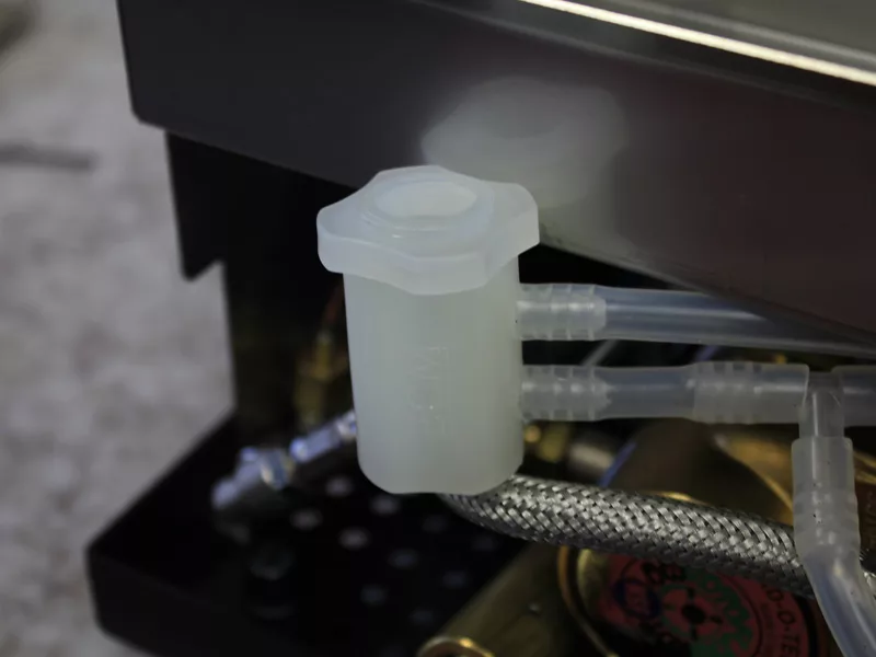 Close-up of a translucent plastic container, likely part of a water tank manifold, with attached tubes connected to metal hoses and equipment. The background is slightly out of focus. © 2025 1st-line.com