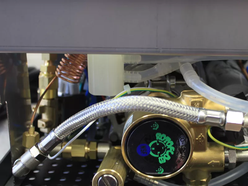 Close-up of internal components of a coffee machine, showing metal pipes, a water tank manifold, a pressure gauge with a green dial, coiled copper tube, and various connectors and wires. © 2025 1st-line.com