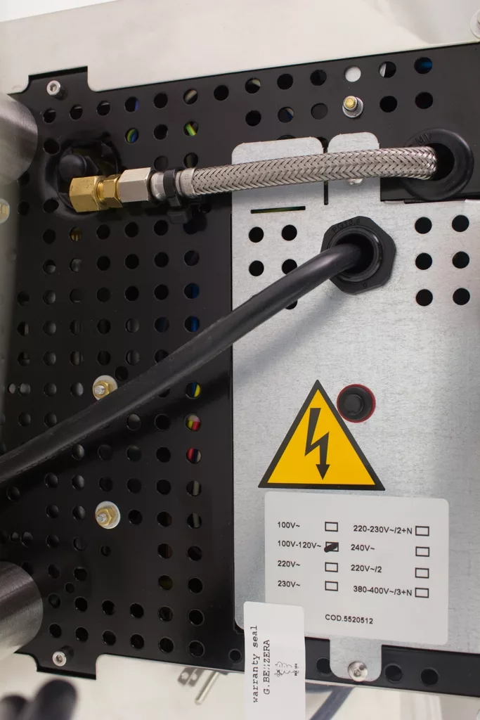 Close-up of the back panel of a Bezzera Mitica V2 HX Espresso Machine, featuring plugged-in cables, a flexible metal conduit, a warning sign with a lightning bolt, and a voltage label. © 2025 1st-line.com