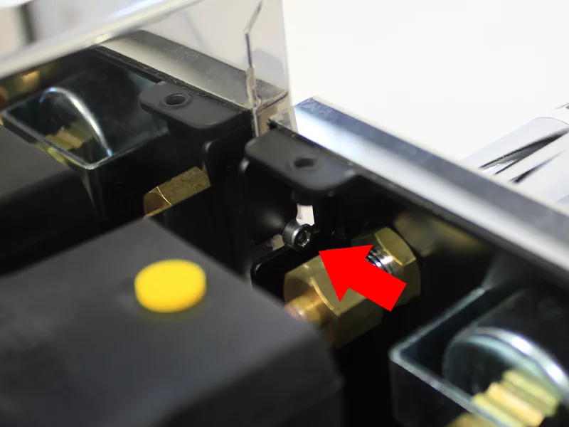 Close-up view of a mechanical component with a red arrow pointing to a screw attached to a black metal bracket—various bolts and connectors for the water tank manifold replacement are visible nearby. © 2025 1st-line.com