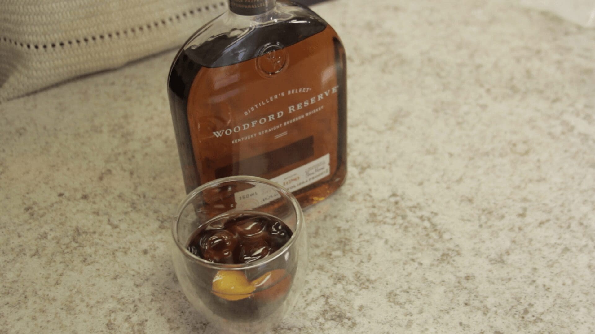 A bottle of Woodford Reserve bourbon sits on a countertop next to a glass filled with ice, dark liquid, and a twist of orange peel—perfect for crafting a classic Cold Fashioned. © 2025 1st-line.com