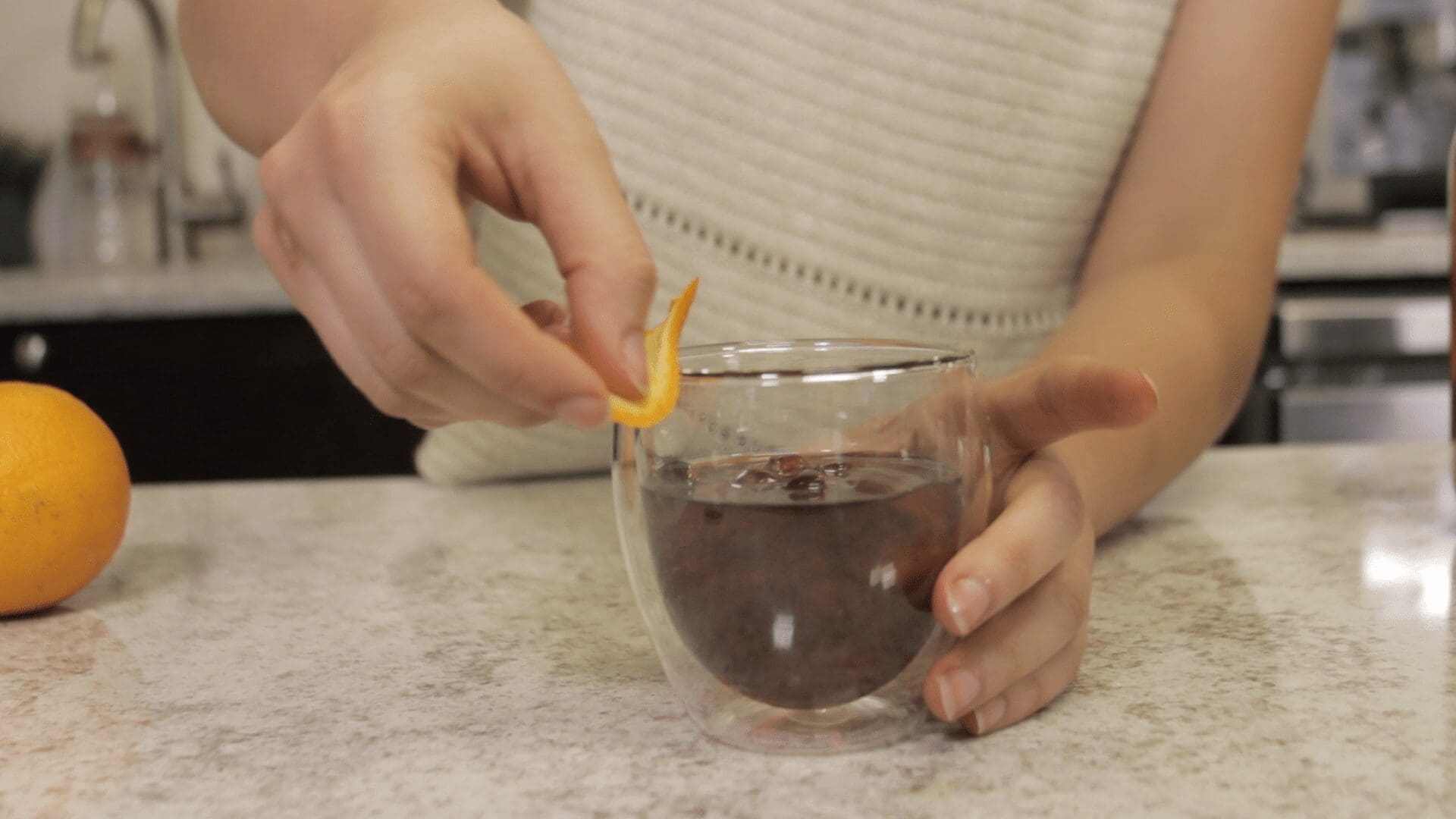 A person holding a glass of dark Cold Fashioned on a countertop, garnishing it with an orange peel. An orange sits to the side on the counter. © 2025 1st-line.com