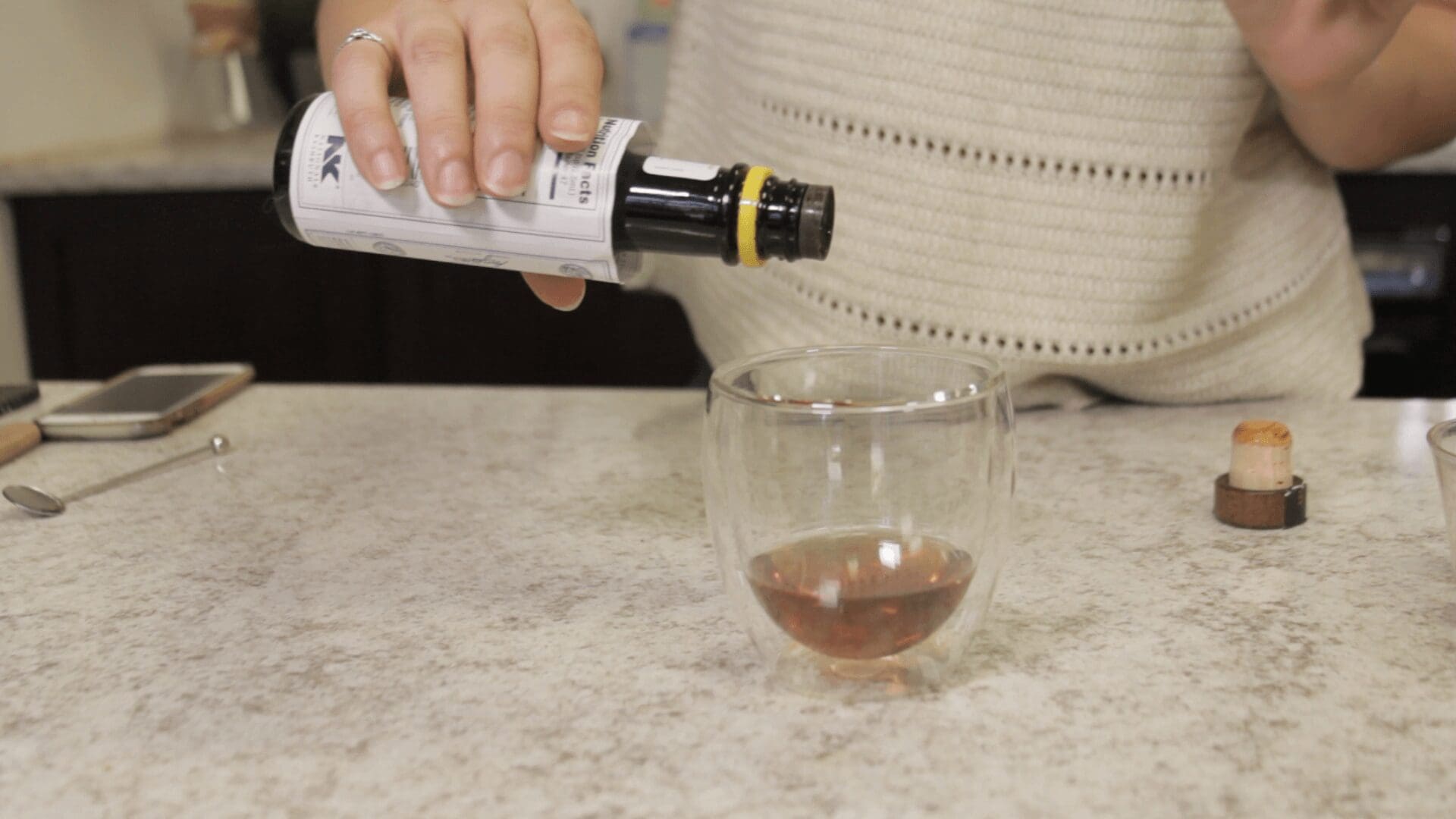 A person pours a Cold Fashioned from a bottle into a clear glass cup on a marble countertop. A phone, spoon, and cork are nearby. Only the persons hand and torso are visible. © 2025 1st-line.com