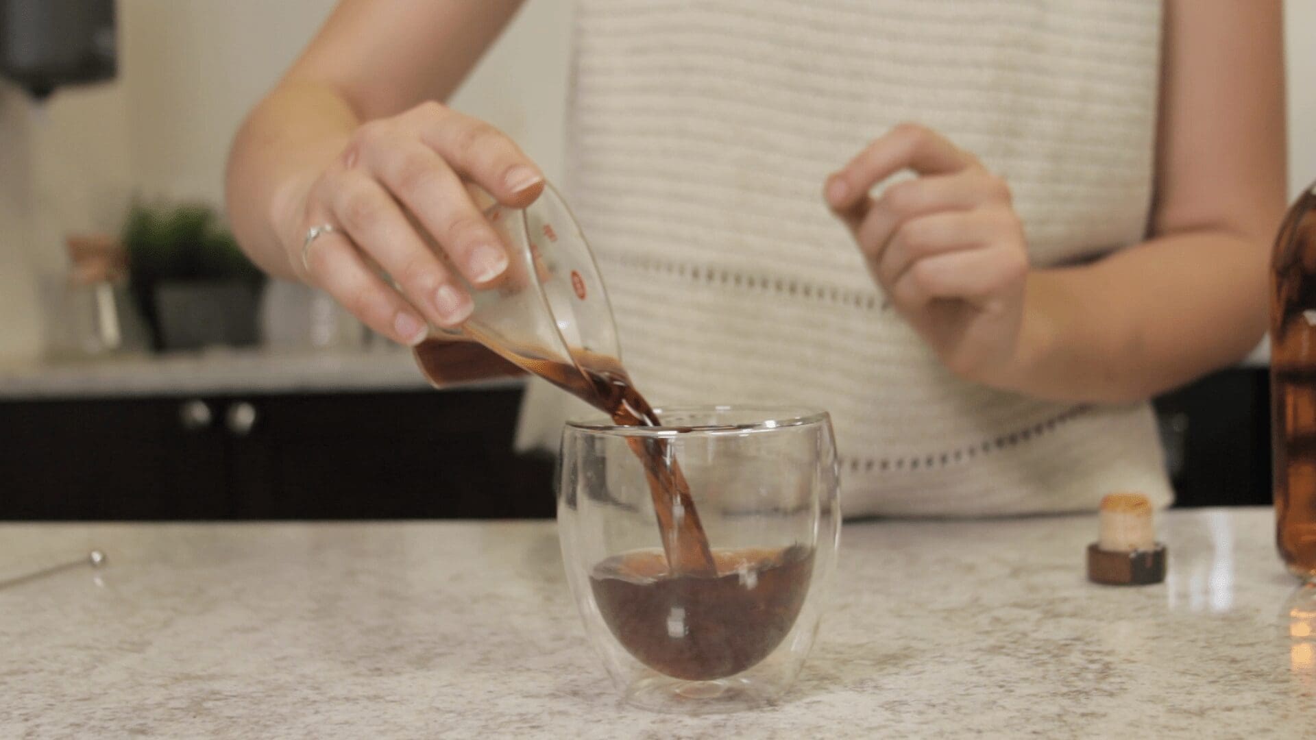A person pours a dark Cold Fashioned from a small measuring cup into a double-walled glass on a kitchen counter. The background features a countertop and blurred kitchen items. © 2025 1st-line.com