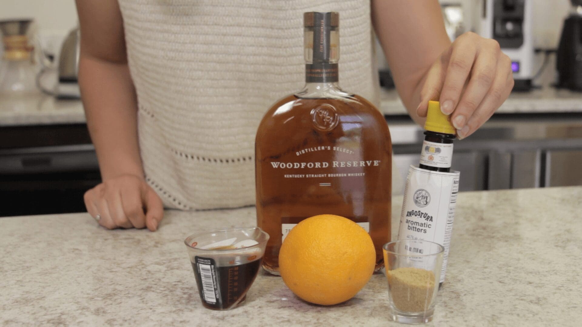 A person stands behind a counter preparing a Cold Fashioned, with a bottle of Woodford Reserve bourbon, angostura aromatic bitters, a cup of liquid, brown sugar, and an orange arranged on the counter. © 2025 1st-line.com