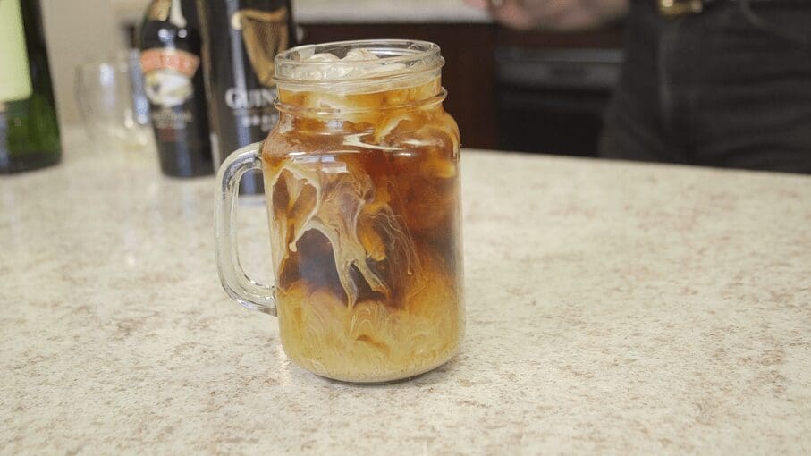 A glass mason jar mug filled with iced coffee, cream swirling through the drink, sits on a light-colored countertop. Bottles of liqueur are blurred in the background, creating a festive St. Patricks Day vibe. © 2025 1st-line.com