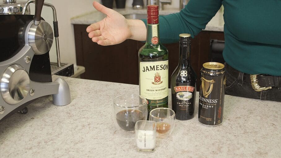 A person gestures toward ingredients for an Irish coffee on a kitchen counter—perfect for St. Patricks Day—including Jameson whiskey, Baileys Irish Cream, Guinness, coffee, cream, and a small glass of brown liquid. © 2025 1st-line.com