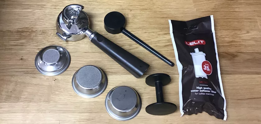 Espresso machine accessories for the Lelit Elizabeth V3 Dual Boiler Espresso Machine on a wooden surface, including a portafilter, filter baskets, plastic tamper, black dosing tool, and water softener filter packet. © 2025 1st-line.com