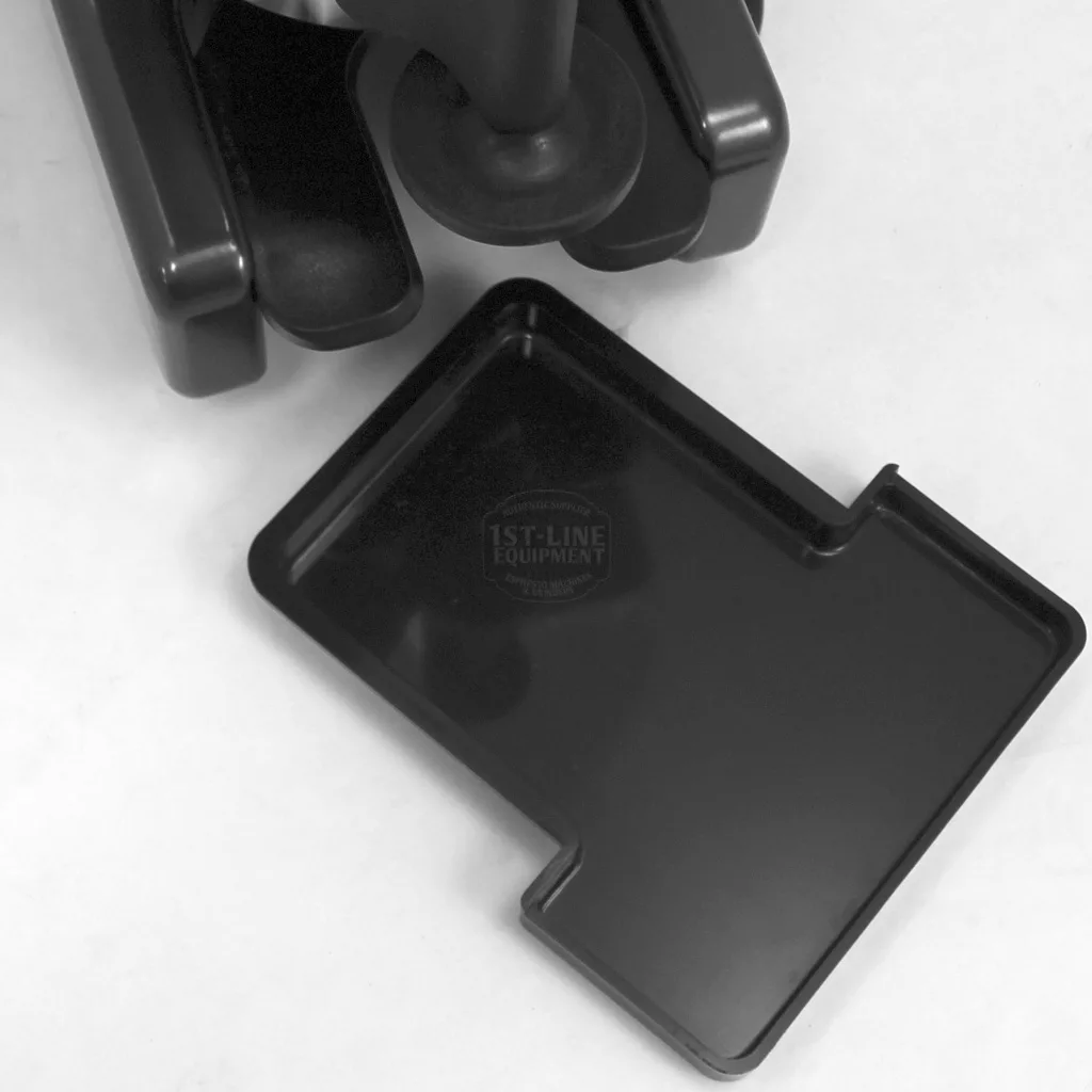 A black plastic rectangular drip tray with a notched edge is displayed on a white surface next to the base of the 1st-line Michael Espresso Coffee Grinder, shown separately from the grinder. © 2025 1st-line.com