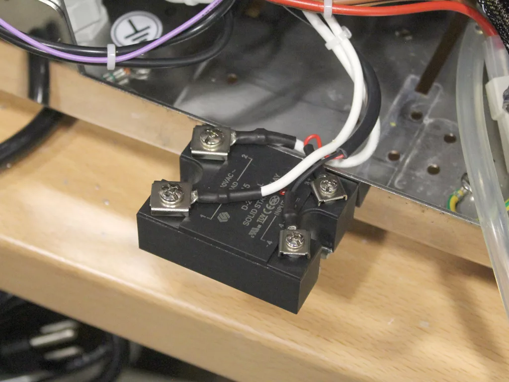 A black solid-state relay, part of a VBM upgrade, is mounted on a metallic surface with red, white, and black wires secured to its terminals by screws. Other electronic components and cables are visible in the background. © 2025 1st-line.com
