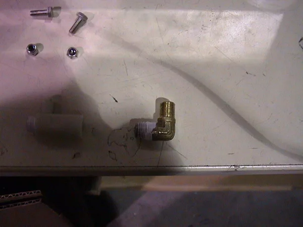 A brass elbow pipe fitting, often used in valve repair or pump repair, is connected to a white plastic threaded pipe on a scratched beige surface, with metal screws and another small white part nearby. © 2025 1st-line.com