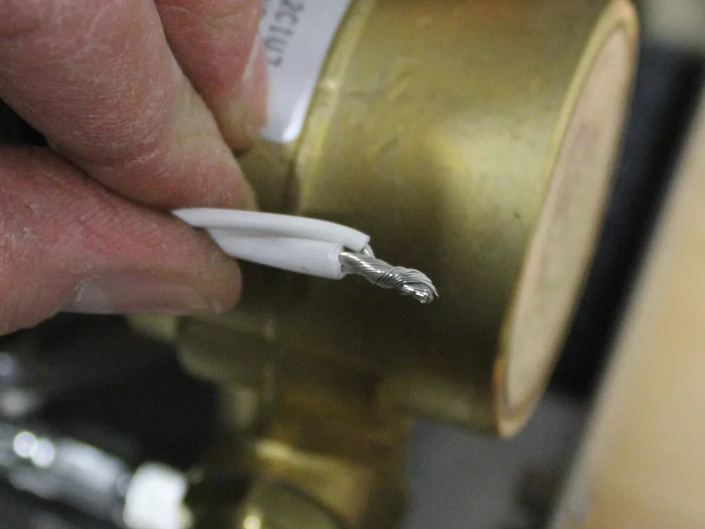 A close-up of a hand holding a piece of white electrical wire with exposed twisted metal strands in front of a brass fitting, commonly seen during a 3.5 to 4.0 conversion on a VBM Dual Boiler espresso machine. © 2025 1st-line.com
