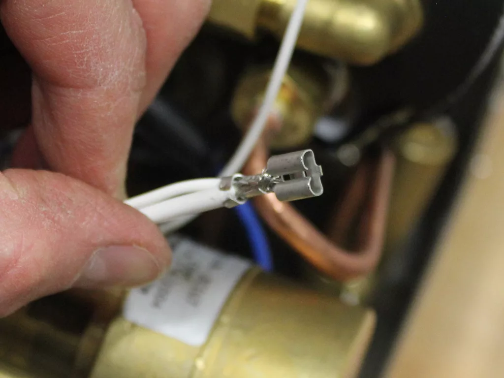 A close-up of a hand holding two white electrical wires with metal connectors attached, positioned near the brass and copper components of a VBM Dual Boiler during a boiler upgrade. © 2025 1st-line.com