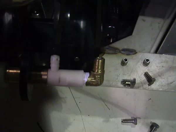A close-up of a mechanical assembly featuring a brass elbow fitting connected to a white plastic pipe, with several loose screws and metal parts scattered on a white surface nearby, typically seen during valve repair. © 2025 1st-line.com