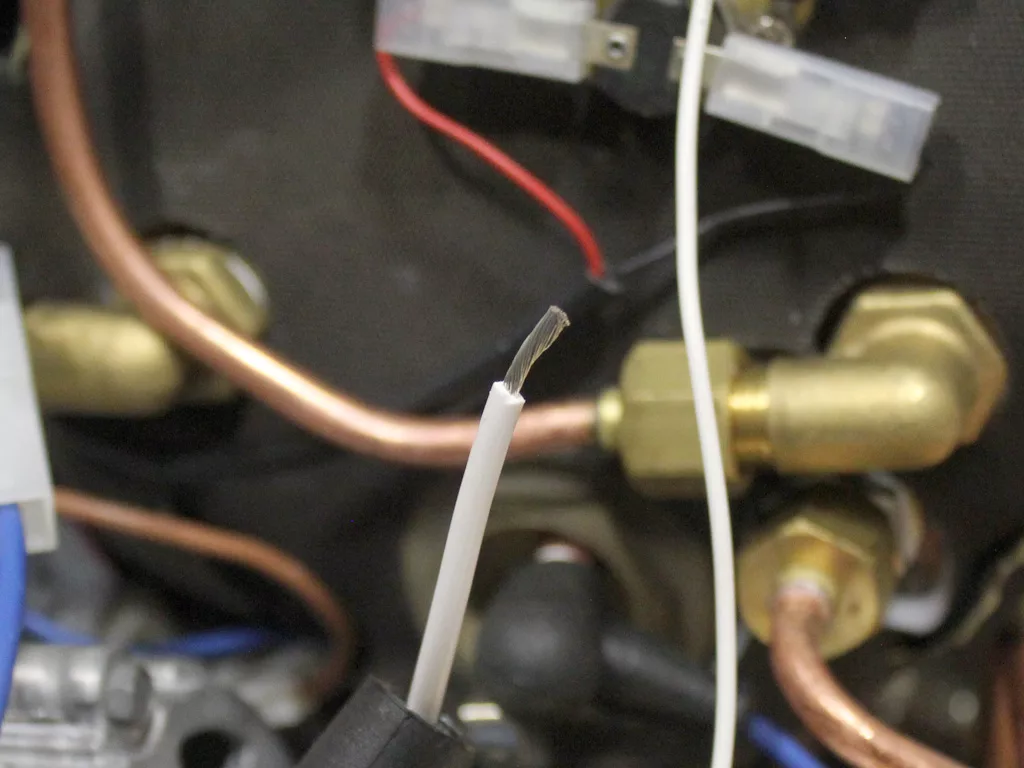 A close-up of a stripped white electrical wire in front of brass fittings and copper pipes, with other wires and connectors visible in the background—ideal for a VBM Dual Boiler 3.5 to 4.0 conversion project. © 2025 1st-line.com