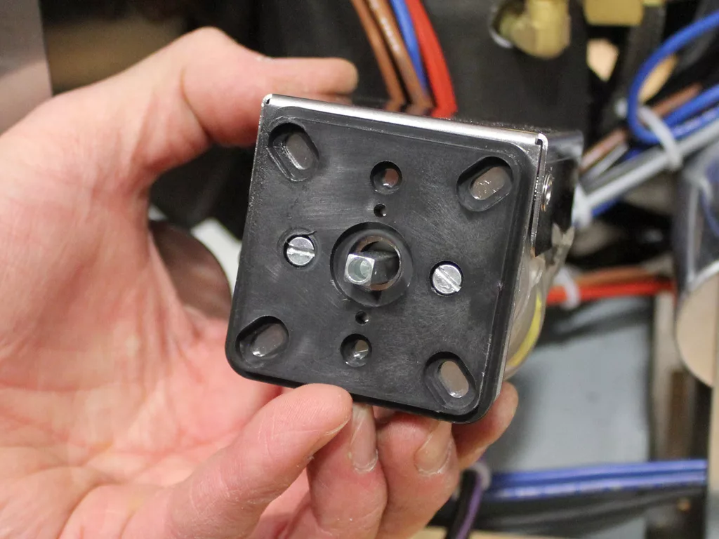 A hand holds a black, square-shaped mechanical part with several holes and metal components, likely part of an espresso machine upgrade or VBM Dual Boiler convert 3.5 to 4.0, against a backdrop of wires and machine parts. © 2025 1st-line.com