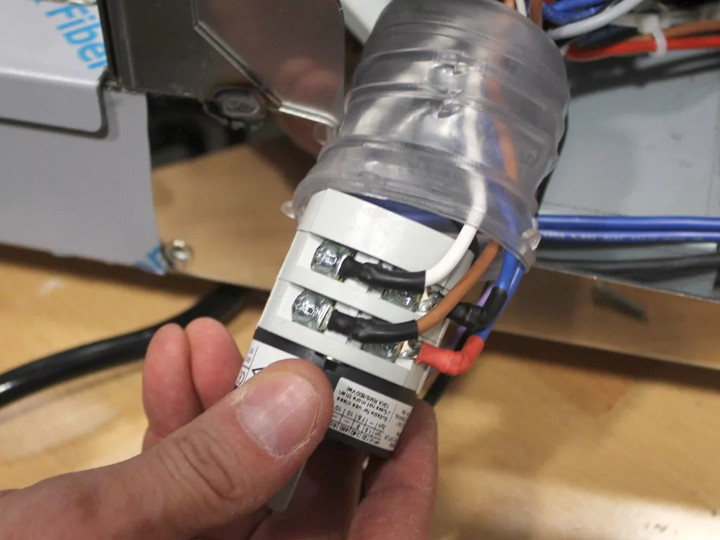 A hand holds an electrical component with exposed wiring and terminals, showing black, brown, blue, and red wires connected to screws during a VBM Dual Boiler conversion inside an open electrical device. © 2025 1st-line.com
