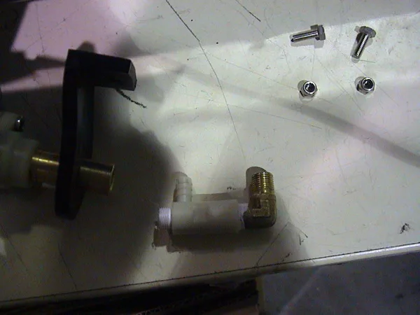 A metal and plastic plumbing fitting, bolts, and washers—used for valve repair—are lying on a scratched white surface beside a black plastic component. © 2025 1st-line.com