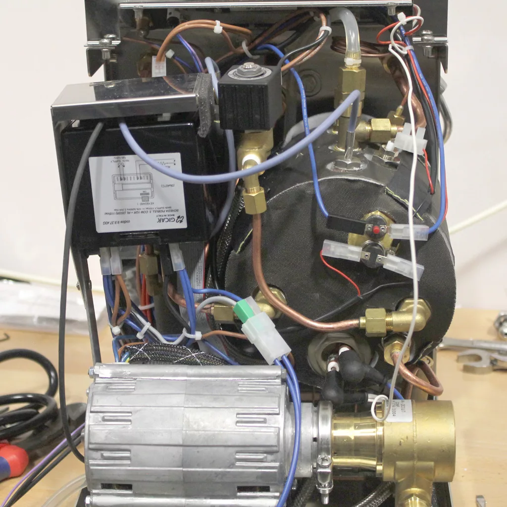 An open VBM Dual Boiler espresso machine reveals internal components, including wires, tubes, a metal motor, brass fittings, a black cylindrical boiler, and various connectors on a wooden surface. © 2025 1st-line.com