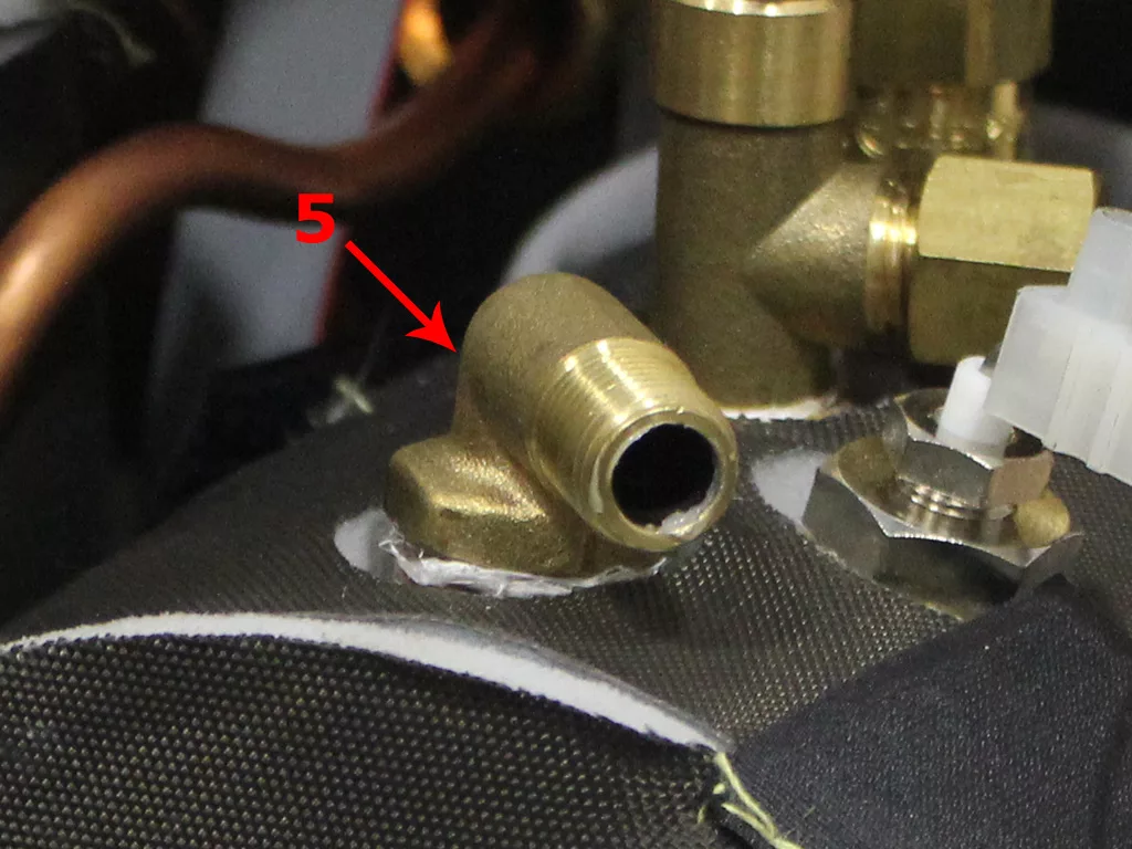 Close-up of a brass pipe fitting with an open end, labeled with a red arrow and the number 5, surrounded by other metal components and black fabric-like material from a VBM Dual Boiler during boiler conversion. © 2025 1st-line.com