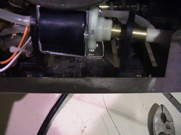 Close-up of a mechanical component with wires attached, possibly part of a machine or appliance. A pair of pliers and a black cable lie on the white surface below, suggesting ongoing pump repair or air release valve repair work. © 2025 1st-line.com