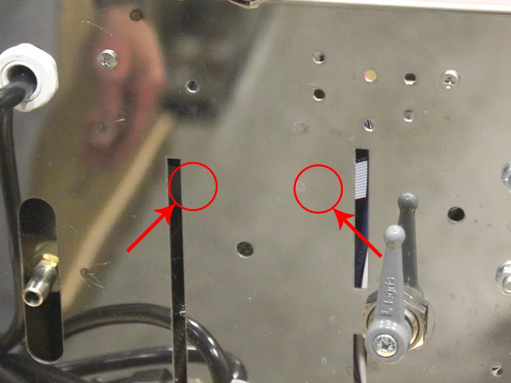Close-up of a metal panel on a VBM Dual Boiler with two red arrows pointing to small holes, a gray lever on the right, black cables below, and a faint reflection of a hand in the background—ideal for converting 3.5 to 4.0. © 2025 1st-line.com
