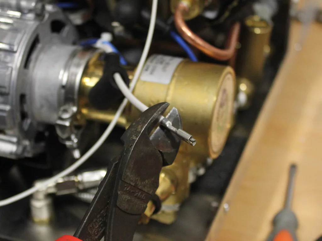 Close-up of a pair of wire cutters gripping a small metal rod, with machinery, VBM Dual Boiler components, and wires visible in the background. © 2025 1st-line.com