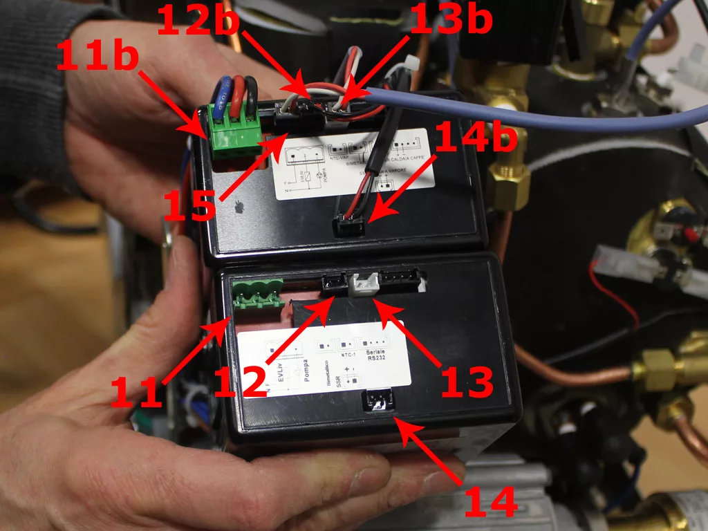 Close-up of a person’s hands holding two black electronic modules, such as those found in a VBM Dual Boiler, connected by wires, with red arrows and labels (11, 11b, 12, 12b) indicating connectors for a 3.5 to 4.0 conversion. © 2025 1st-line.com