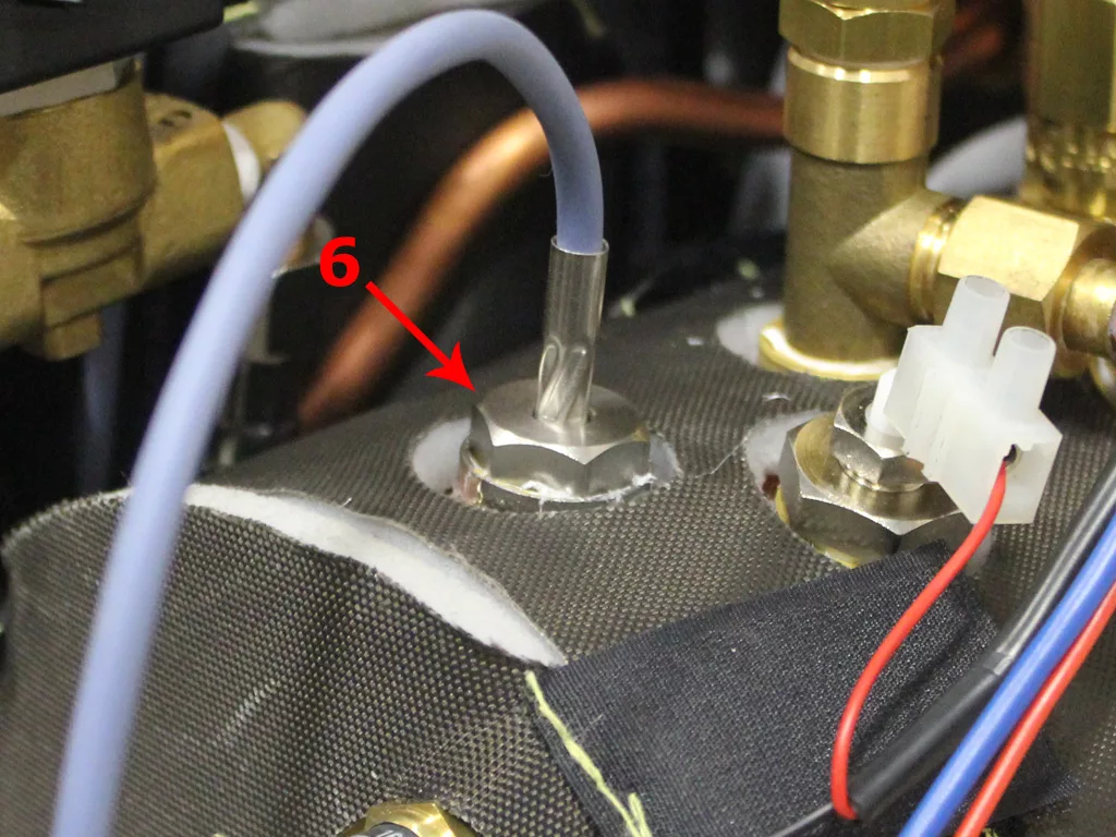 Close-up of a VBM Dual Boiler espresso machine component with pipes and wiring, showing a metal fitting labeled with a red arrow and the number 6, attached to a metallic surface surrounded by other connectors. © 2025 1st-line.com